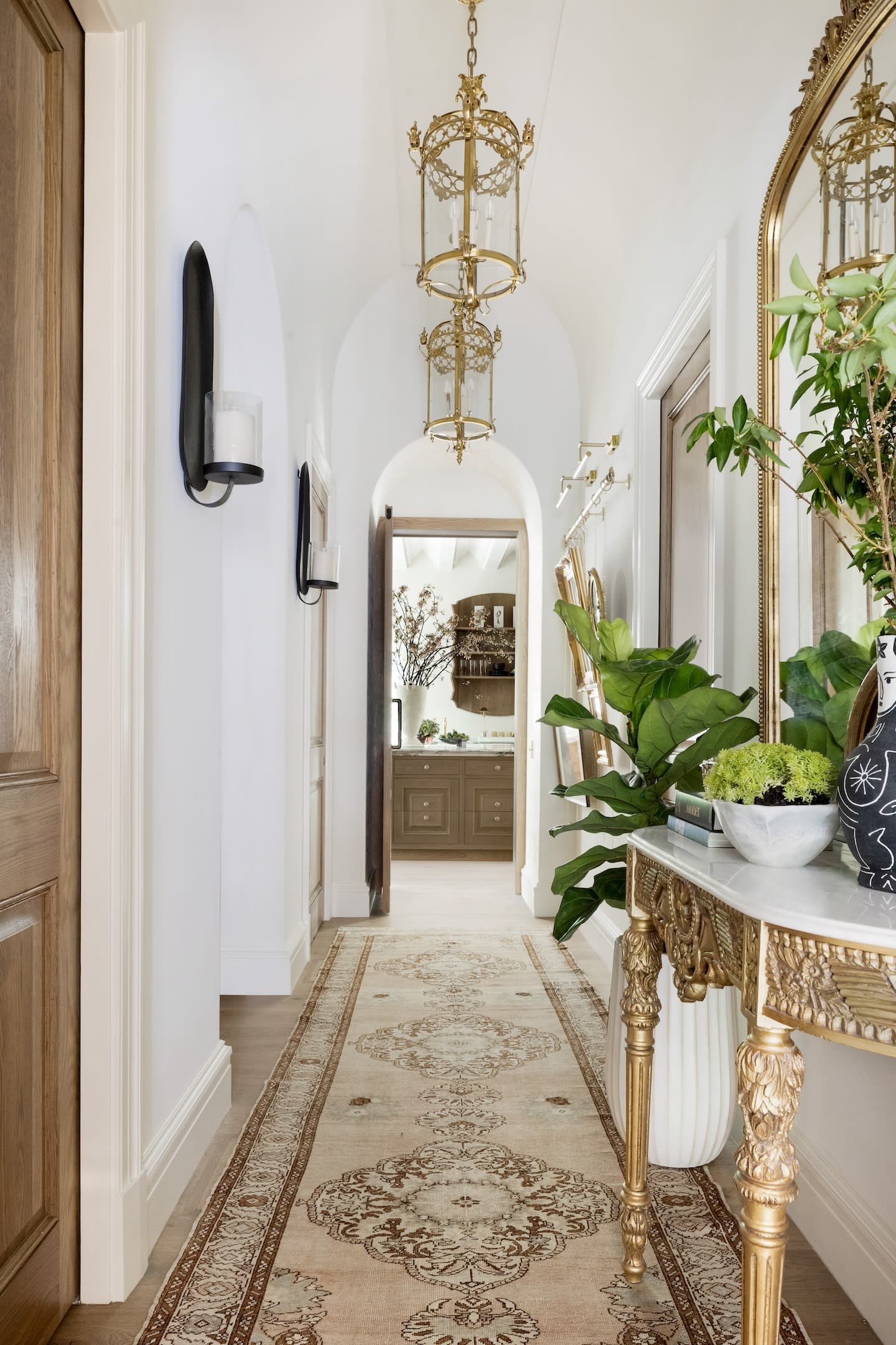 Elegant hallway with a long, ornate rug, gold-accented furniture, large mirror, and lush greenery. Warm lighting creates a welcoming atmosphere.